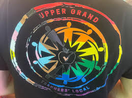 A photo of the back of a tshirt with the logo of Upper Grand Elementary Teachers' Federation of Ontario superimposed on the back overtop of the Progress Pride flag